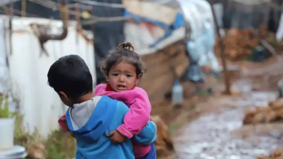 A boy carries his sister through a refugee village wearing his fathers too large shoes